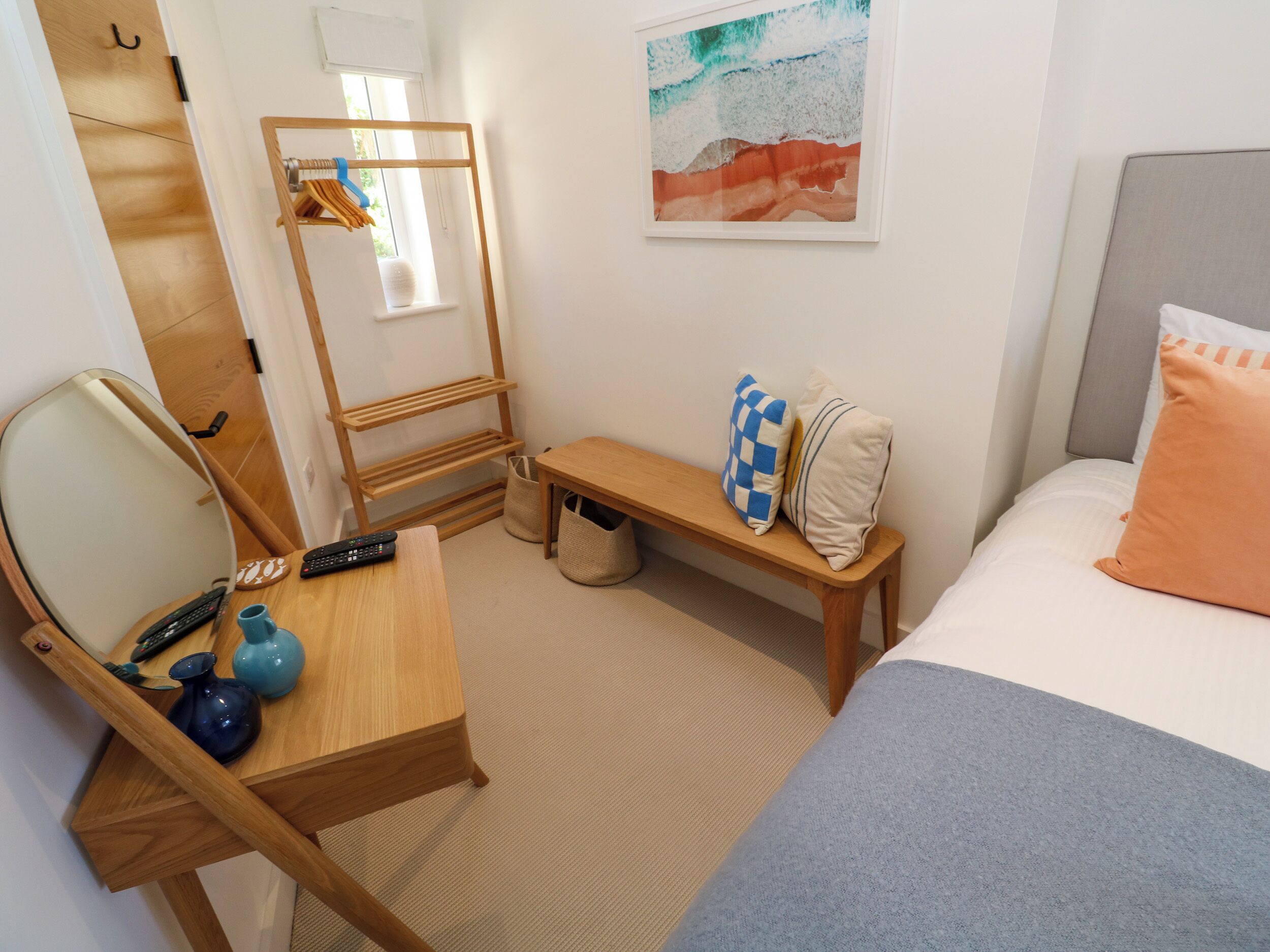 A hotel room with a bed, a wooden desk with a mirror, a chair, a small table, a bench, and a wall-mounted rack with clothes.