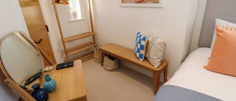A hotel room with a bed, a wooden desk with a mirror, a chair, a small table, a bench, and a wall-mounted rack with clothes.