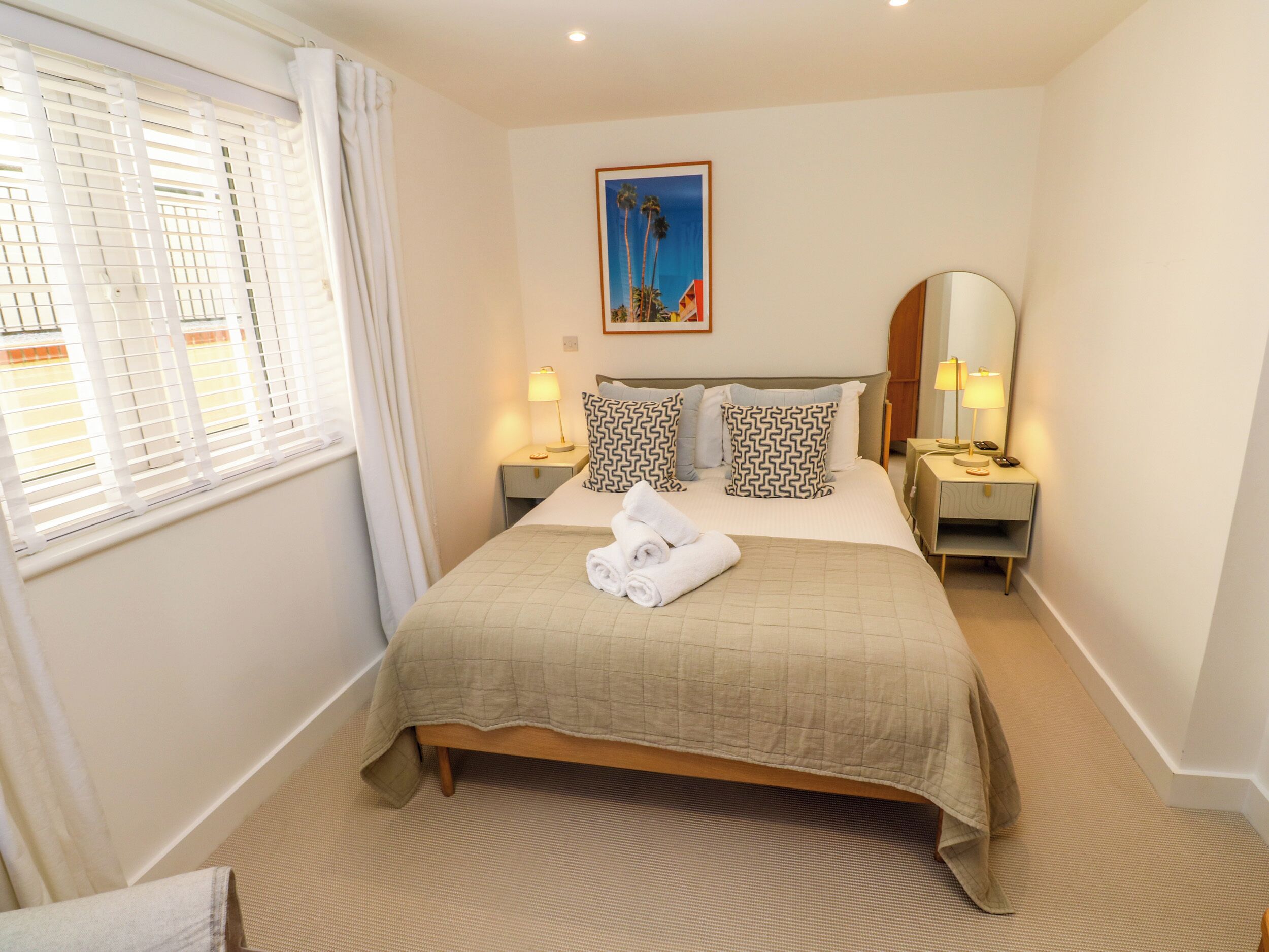 A neatly arranged hotel room with a bed, two bedside tables, a nightstand, a window with blinds, a painting on the wall, and a mirror on the nightstand.