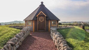 Een vakantiehuisje van hout met een zeshoekige deur, omgeven door een stenen muur en een grindpad, gelegen in een grasveld met heuvels in de verte.