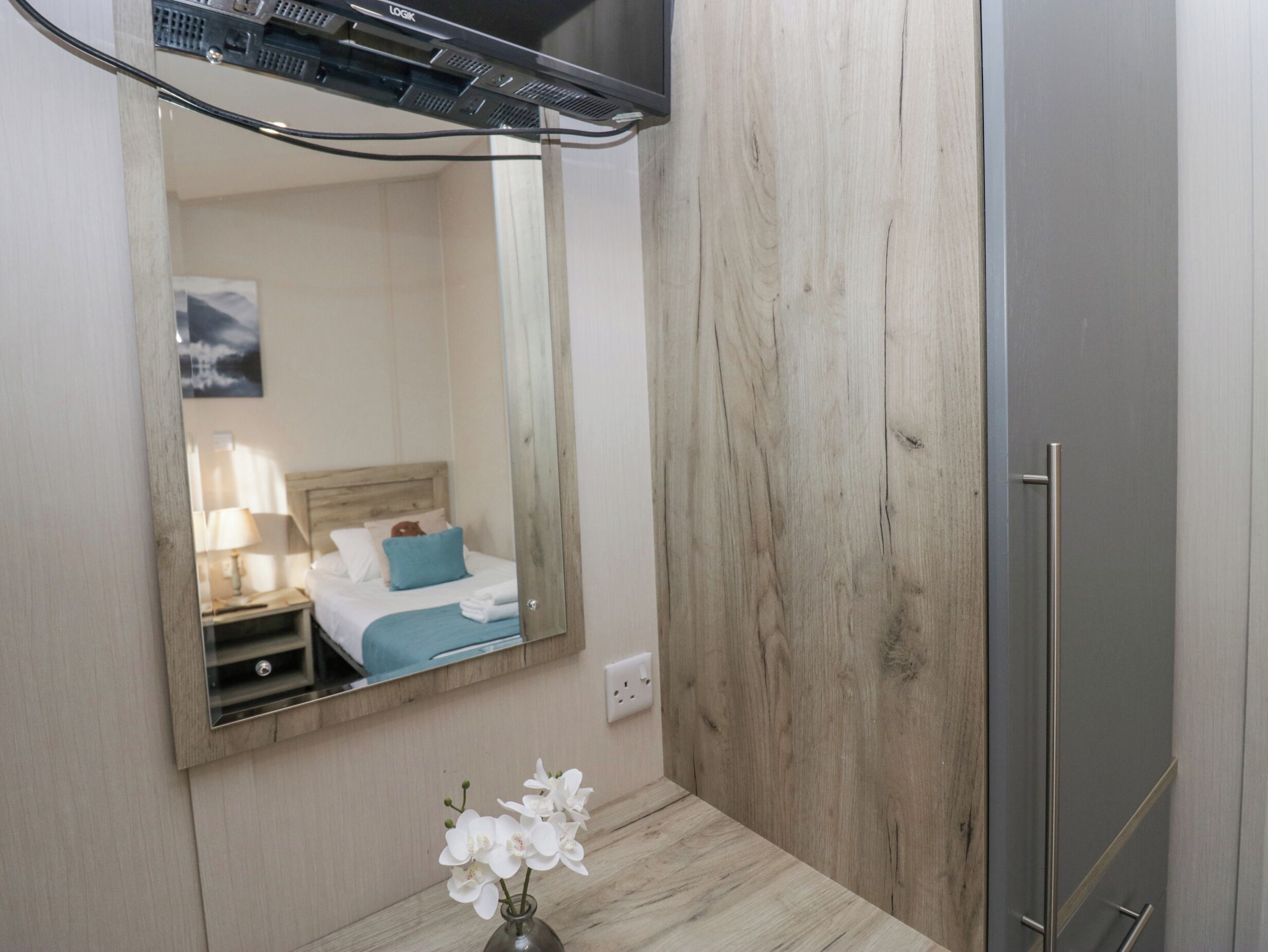 Une chambre d’hôtel avec un mur en bois, un lit avec un oreiller bleu, une table de chevet avec une lampe et un miroir qui reflète la pièce.