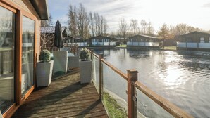 A wooden deck with wicker furniture overlooking a calm water body, with buildings in the background. - The Three Bears Lodge, Coniston Drive 11 (Carnforth)