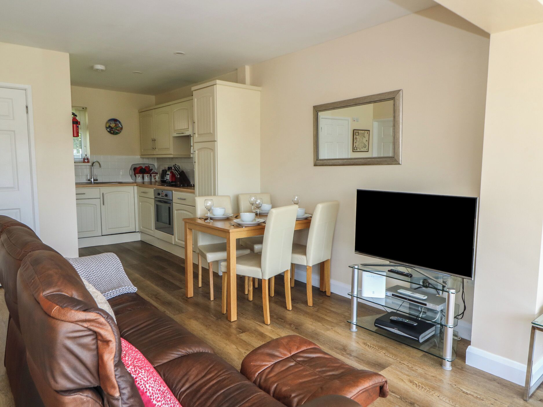 A modern living room with a brown leather sofa, a dining table with chairs, a TV on a glass stand, and a kitchen in the background.