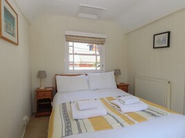 Una cama bien hecha con sábanas blancas y una manta a rayas, dos lámparas de noche, una ventana con cortinero y un cuadro en la pared.