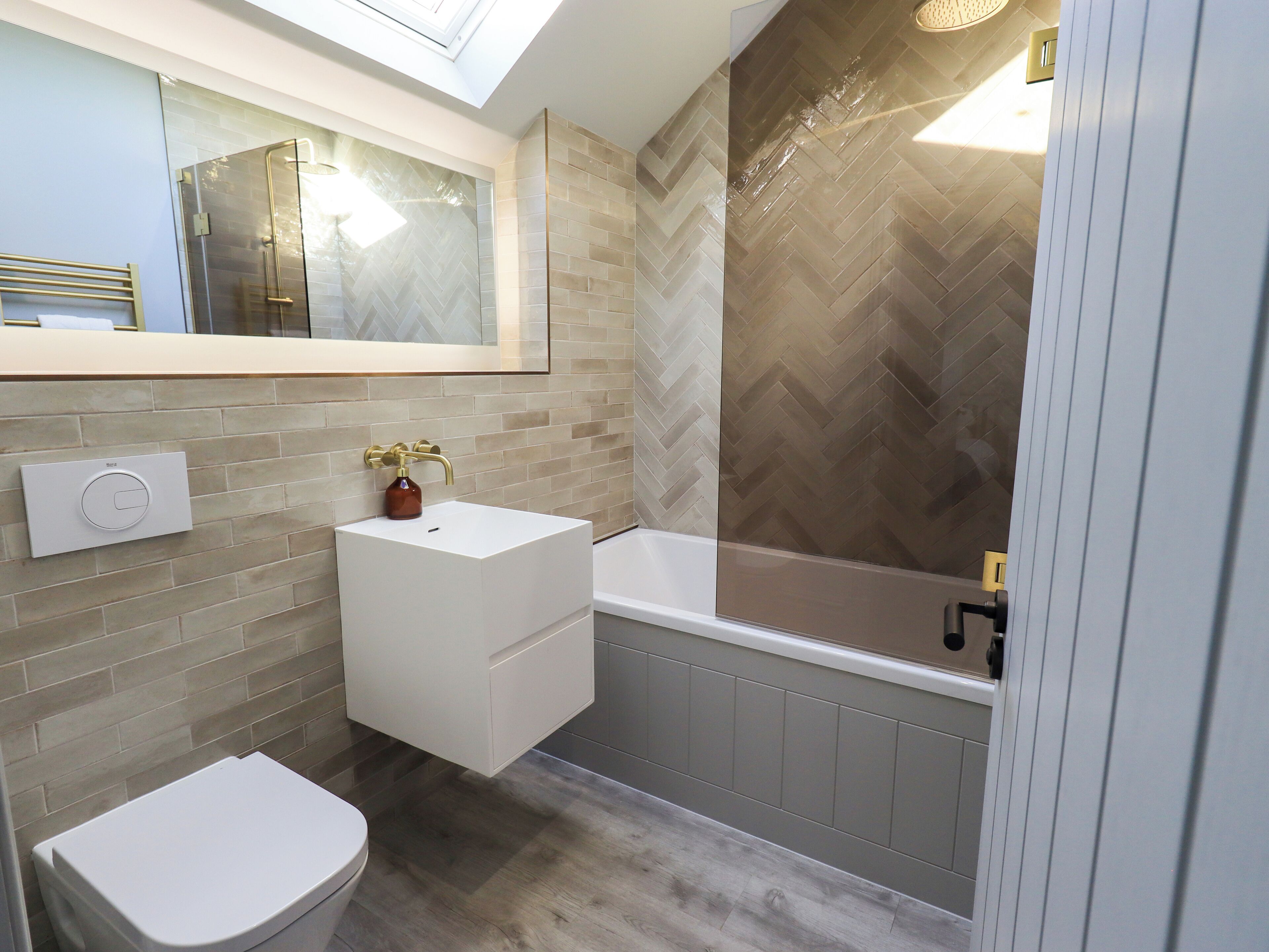 A modern bathroom with a herringbone-patterned wall, a white sink with gold fixtures, a toilet, and a bathtub.