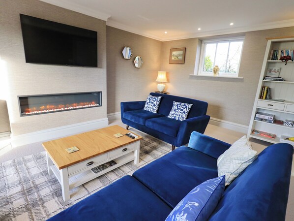 A living room with a flat-screen TV, a fireplace, a blue sofa set, a coffee table, and a bookshelf. - The Elterwater Barn (Workington)