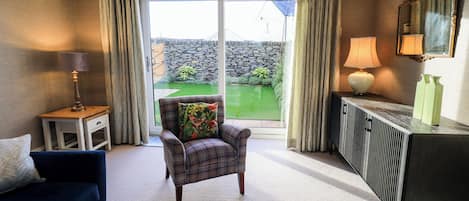 Ein Wohnzimmer mit Sofa, Sessel und Couchtisch, ein Beistelltisch mit Lampe, ein Schrank mit Spiegel und Blick auf einen Garten durch eine Schiebetür.