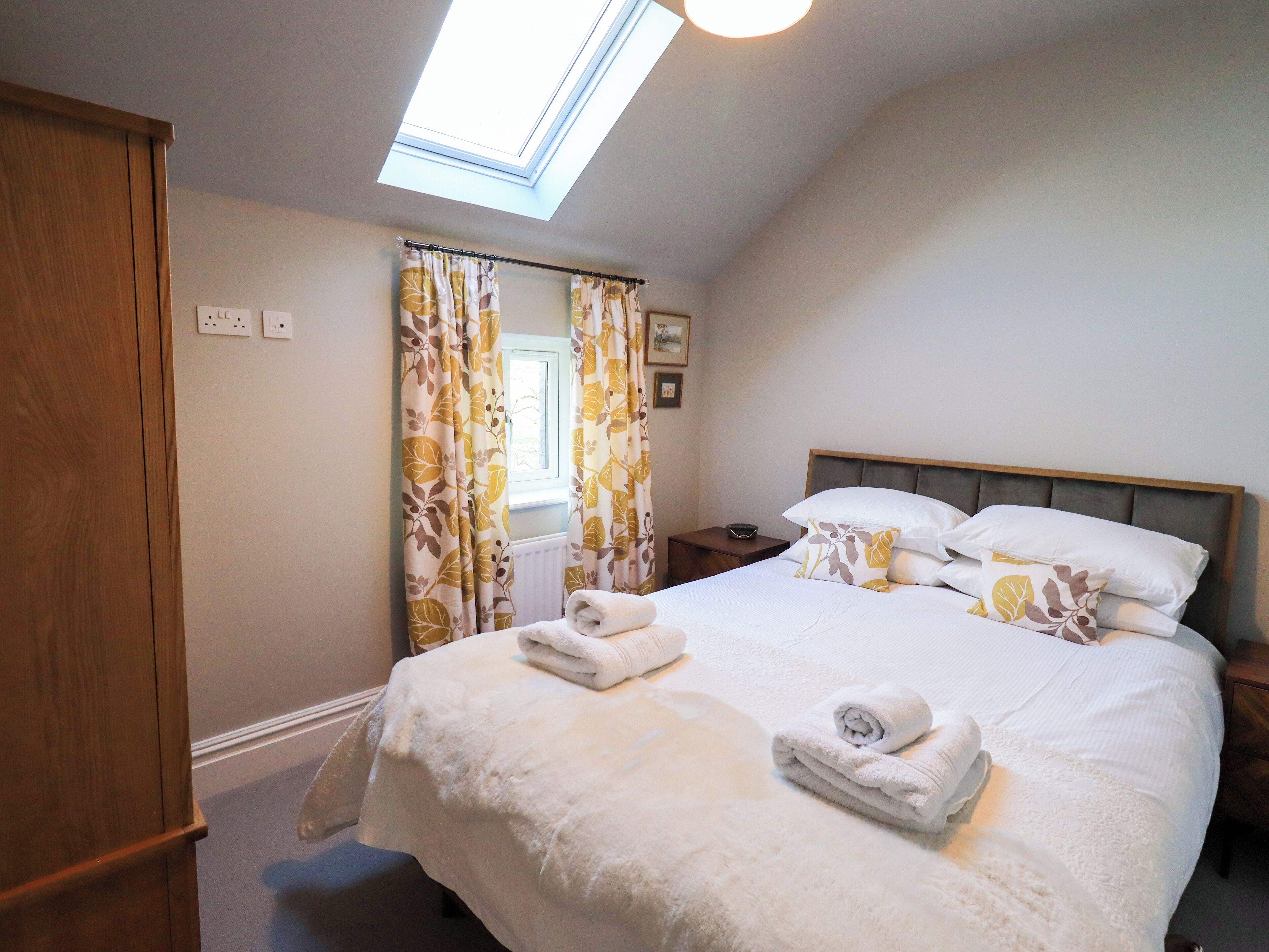 Quarto com claraboia, cama com roupa de cama branca, guarda-roupa de madeira e mesa de cabeceira com uma tigela.
