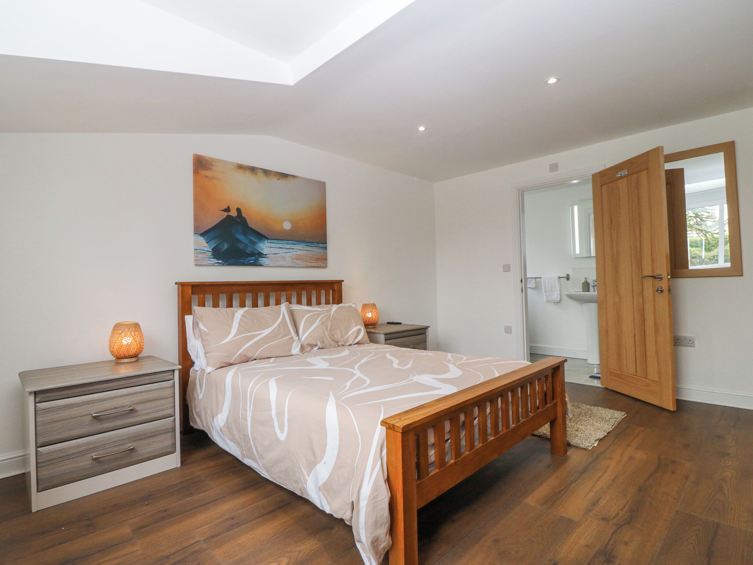 A bedroom with a wooden bed, a nightstand with a lamp, a dresser, a painting of a sunset, and a bathroom visible through an open door.