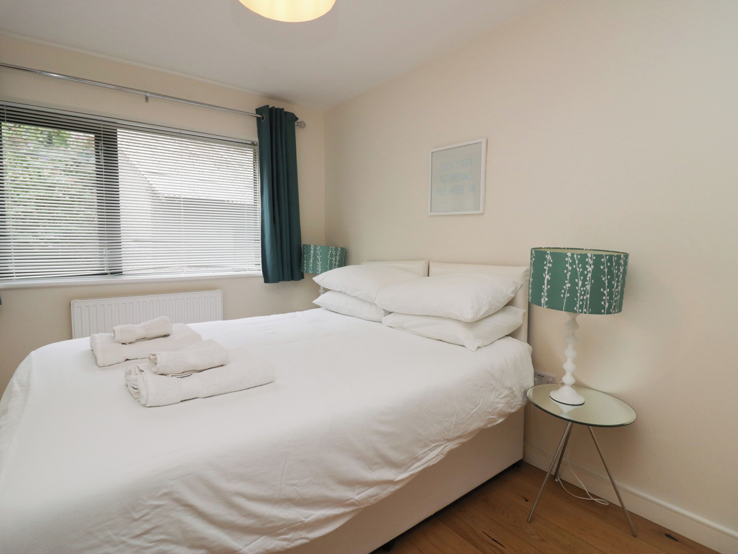A neatly made bed with white linens and towels, a bedside lamp, and a window with blinds and curtains.
