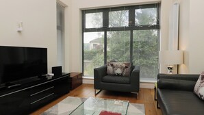 A modern living room with a flat-screen TV, a glass coffee table, a sofa, and a chair with a decorative pillow. - 12 Blue Hill Park (Ambleside)