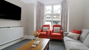 A living room with a flat-screen TV mounted on the wall, a wooden coffee table, two red armchairs, a grey sofa, and a window with curtains. - Park Beck 3 (Windermere)