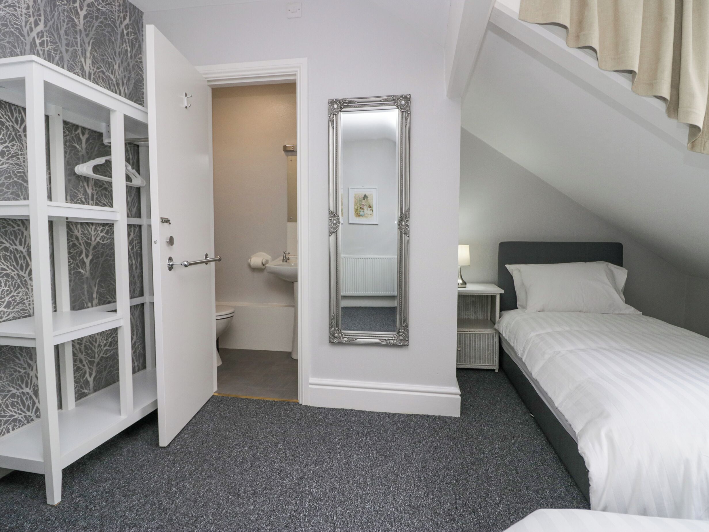 A bedroom with a bed, a nightstand, a dresser, a bathroom with a toilet and sink, and a large mirror.