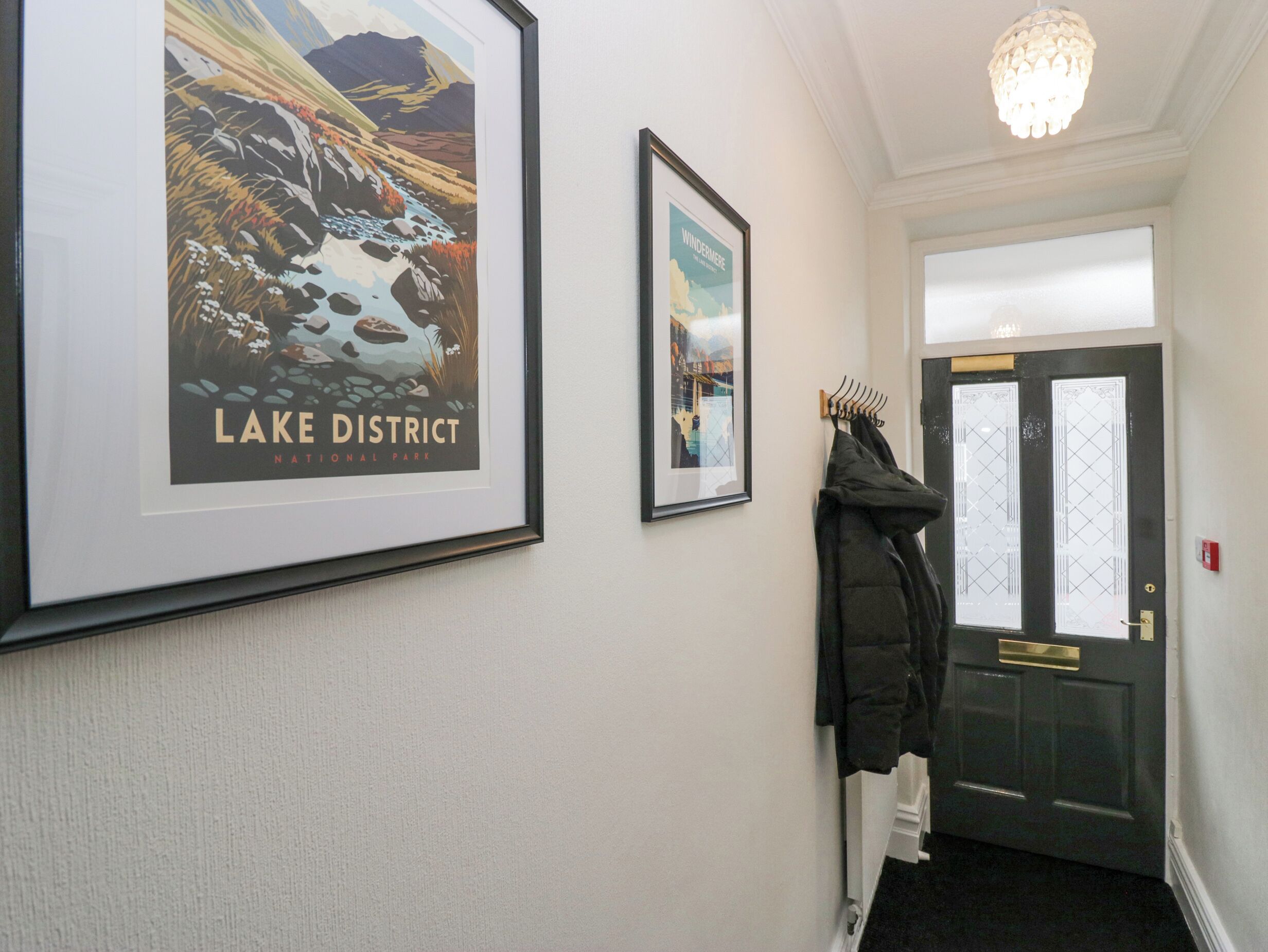 Un couloir orné d’affiches encadrées représentant le parc national du Lake District et le lac de Windermere, un porte-manteau avec une veste, et une porte dotée d’un panneau de verre.