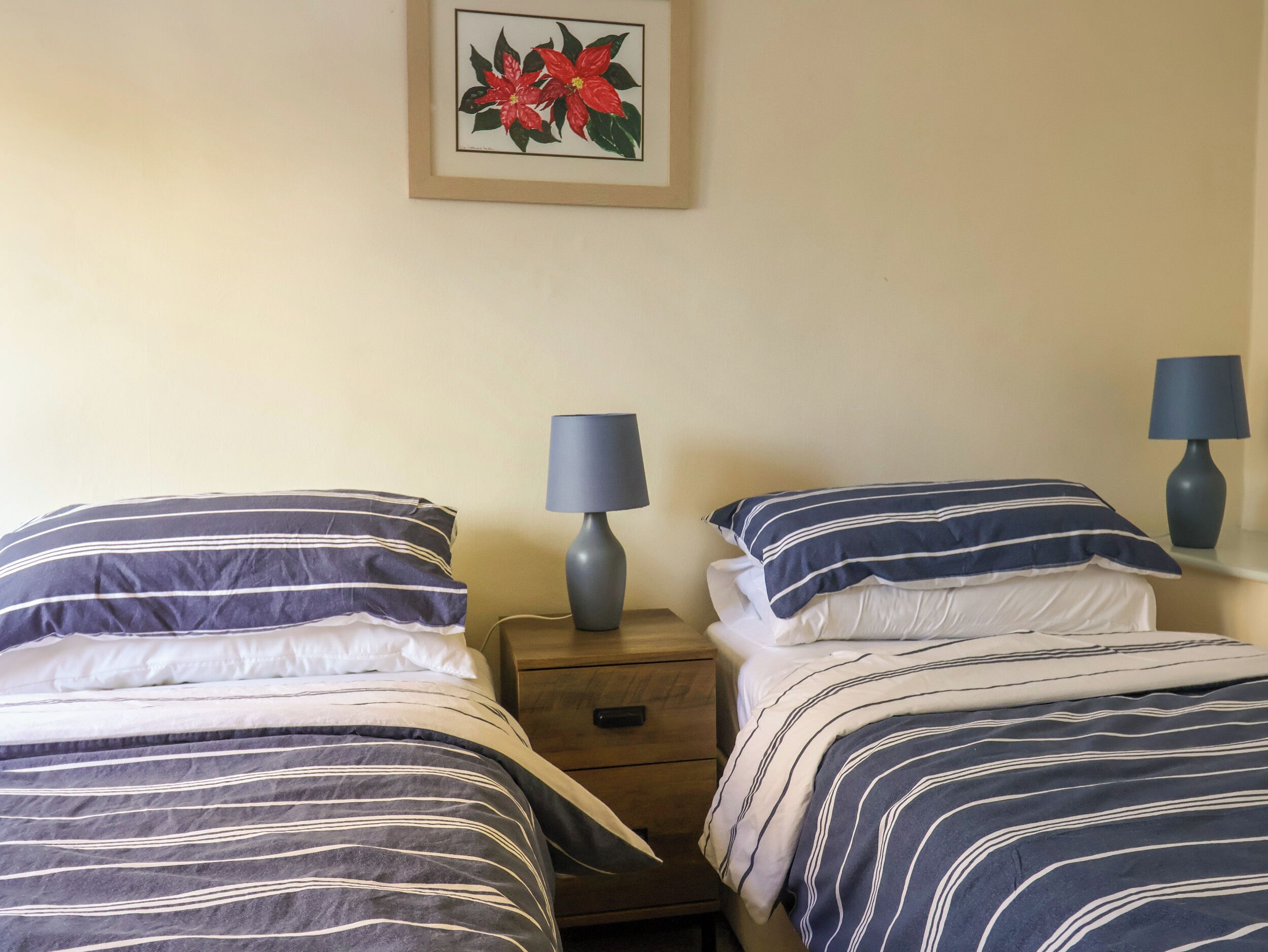A hotel room with two beds, each with striped bedding, a nightstand with a lamp, and a framed picture of red flowers on the wall.