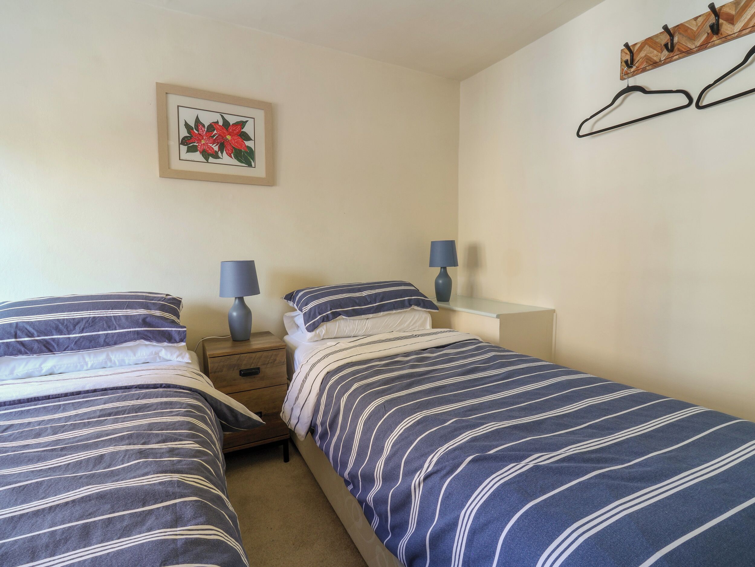 Quarto com duas camas, cada uma com roupa de cama listrada, mesa de cabeceira com abajur, mesa lateral e um quadro na parede.