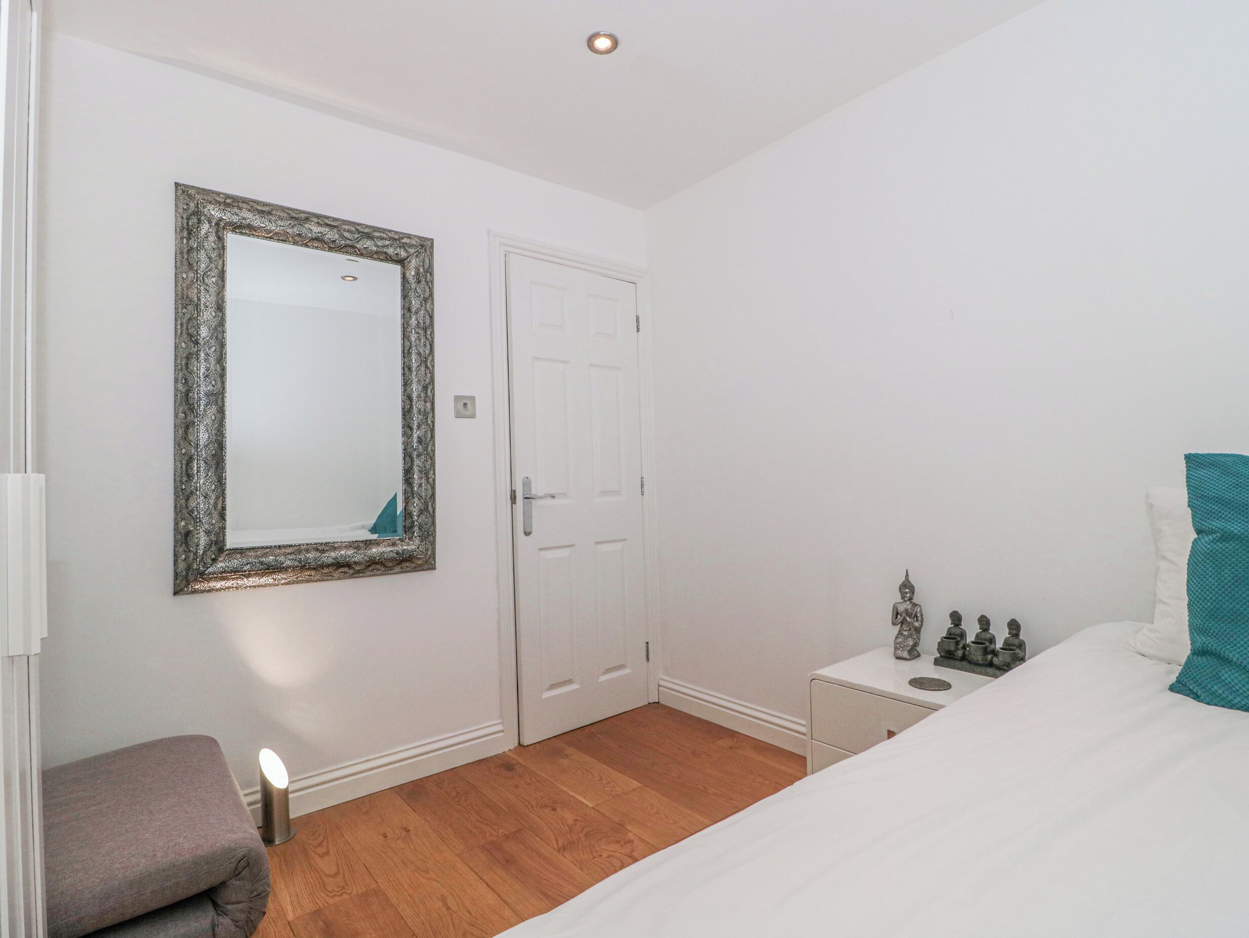 Une chambre à coucher avec un lit, une table de chevet, un petit banc, une porte et un miroir au mur.