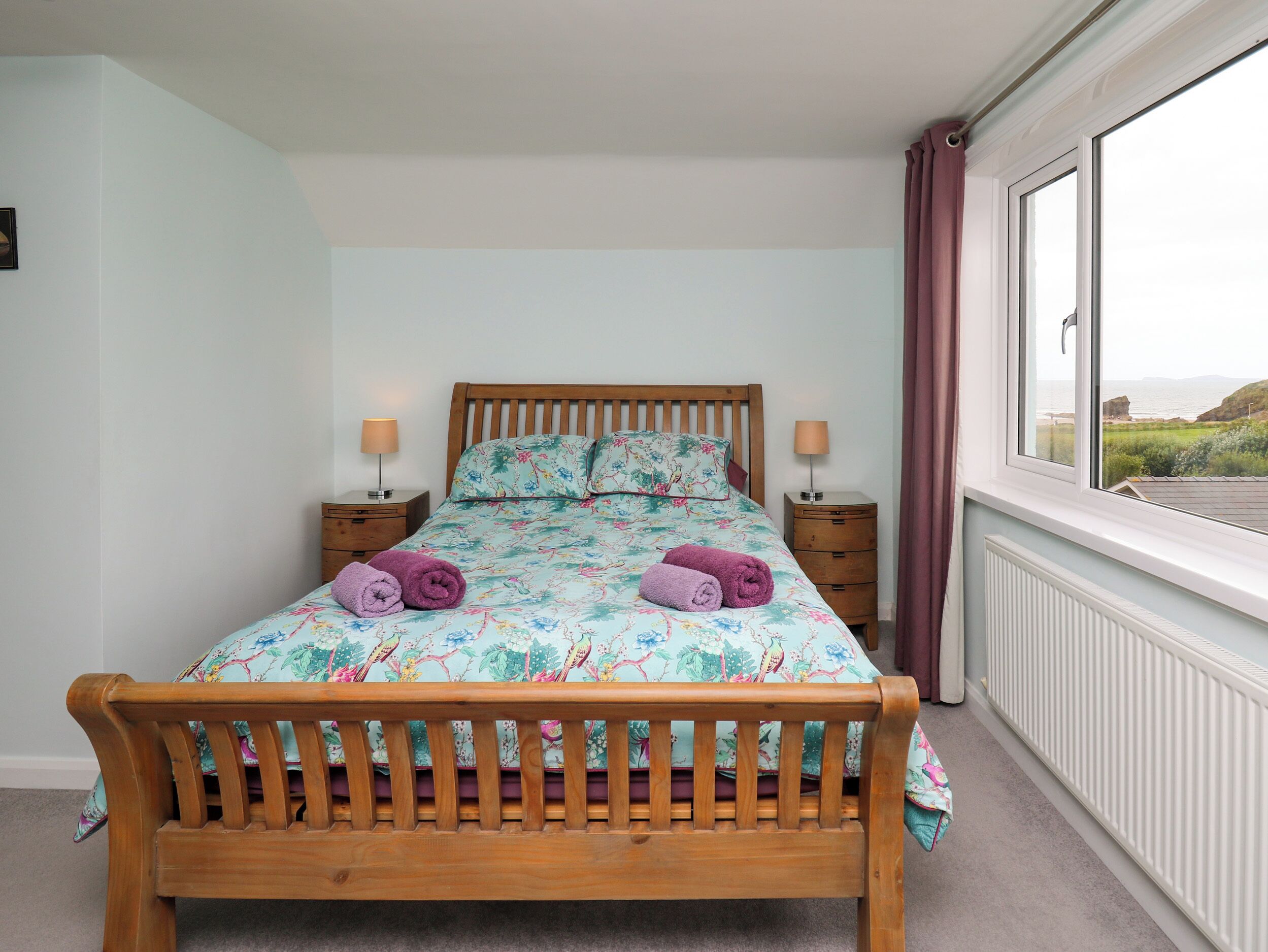 A bedroom with a wooden bed, floral bedding, two bedside tables with lamps, a window with a view of the sea, and a radiator.