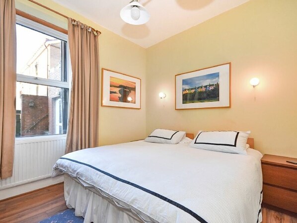 Une chambre à coucher avec un grand lit, deux tables de chevet, une fenêtre avec des rideaux et deux tableaux encadrés accrochés au mur.