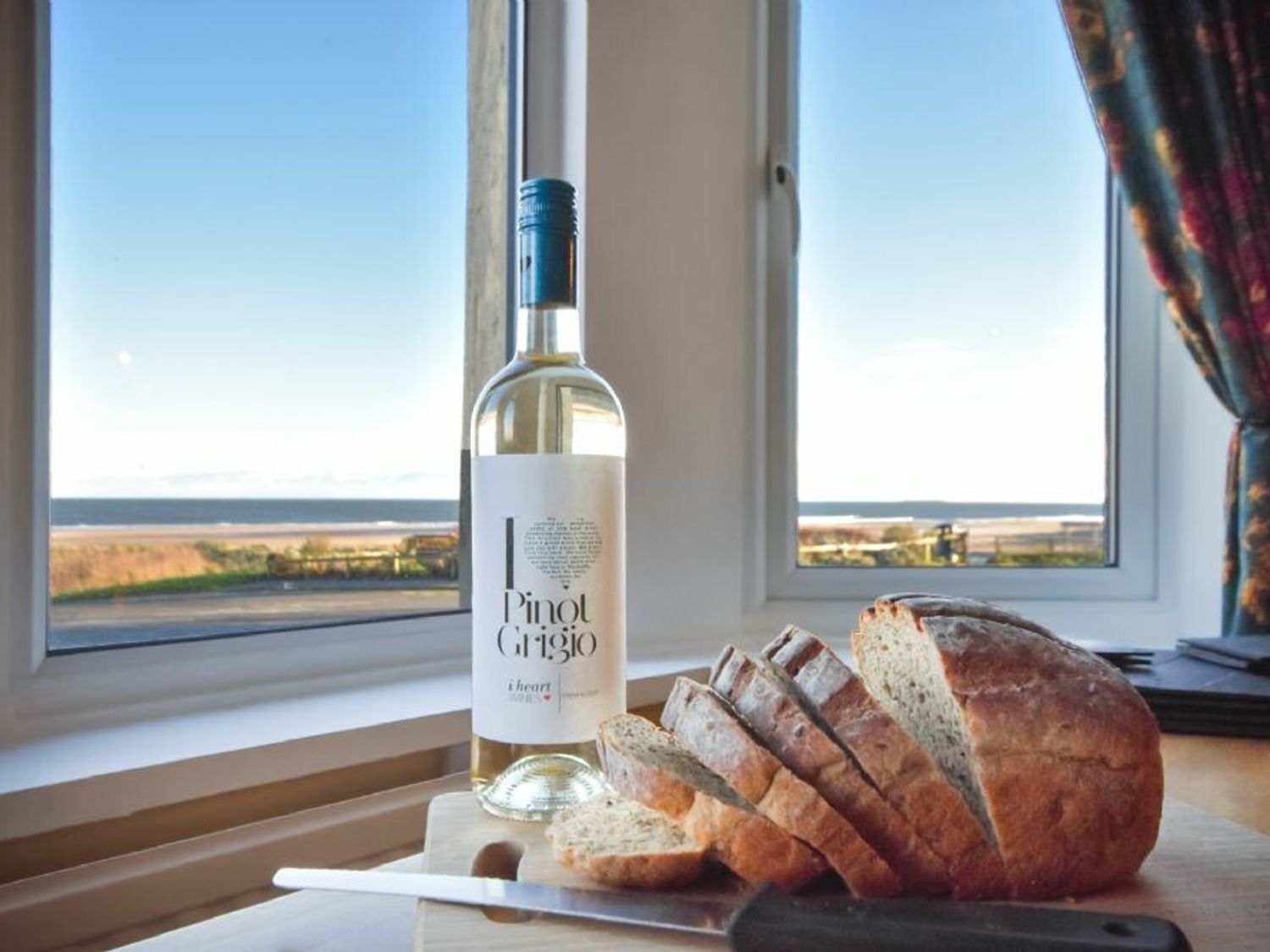 A bottle of Pinot Grigio wine, a loaf of bread, and a knife on a wooden cutting board by a window with a view of the landscape.