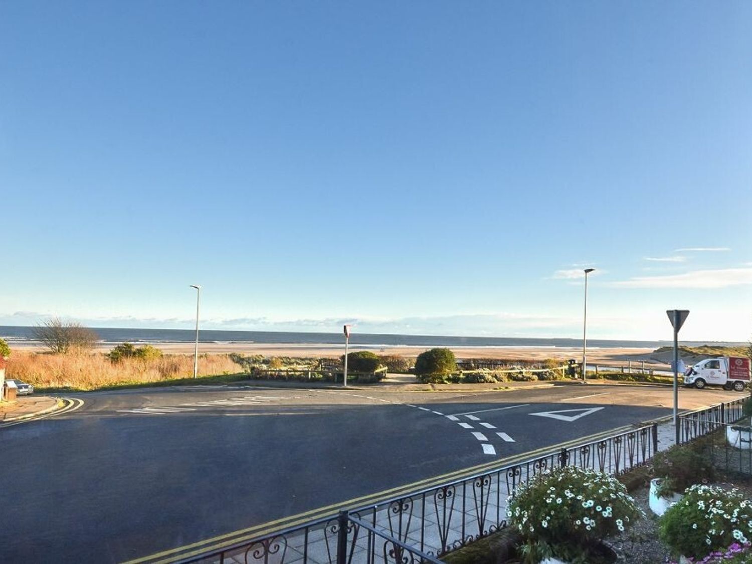 A coastal road with a clear view of the sea, a white van parked on the side, and a metal fence along the road.