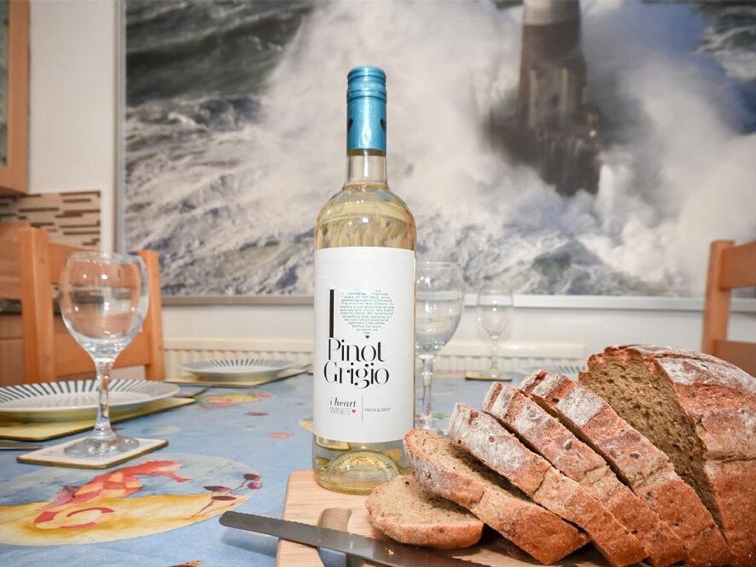 A table with a bottle of Pinot Grigio wine, a loaf of bread, and a painting of a seascape in the background.