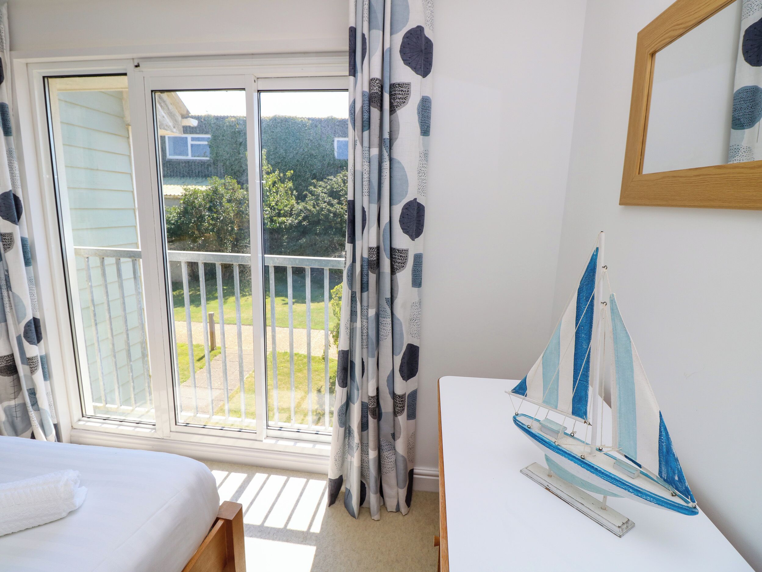A bedroom with a bed, a small table with a sailboat model, a window with curtains, and a view of a garden and house outside.