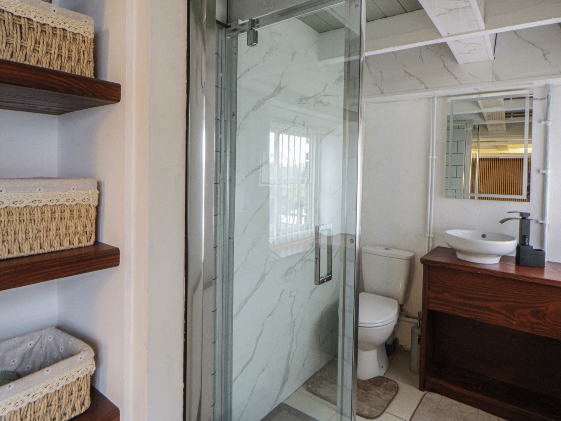 Une salle de bain avec une douche à parois de verre, des toilettes, un lavabo avec un miroir et des paniers en osier placés sur des étagères.