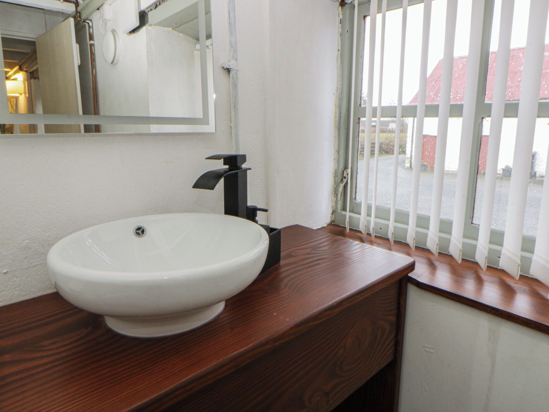 A bathroom with a white sink, wooden vanity, and a window with white blinds.
