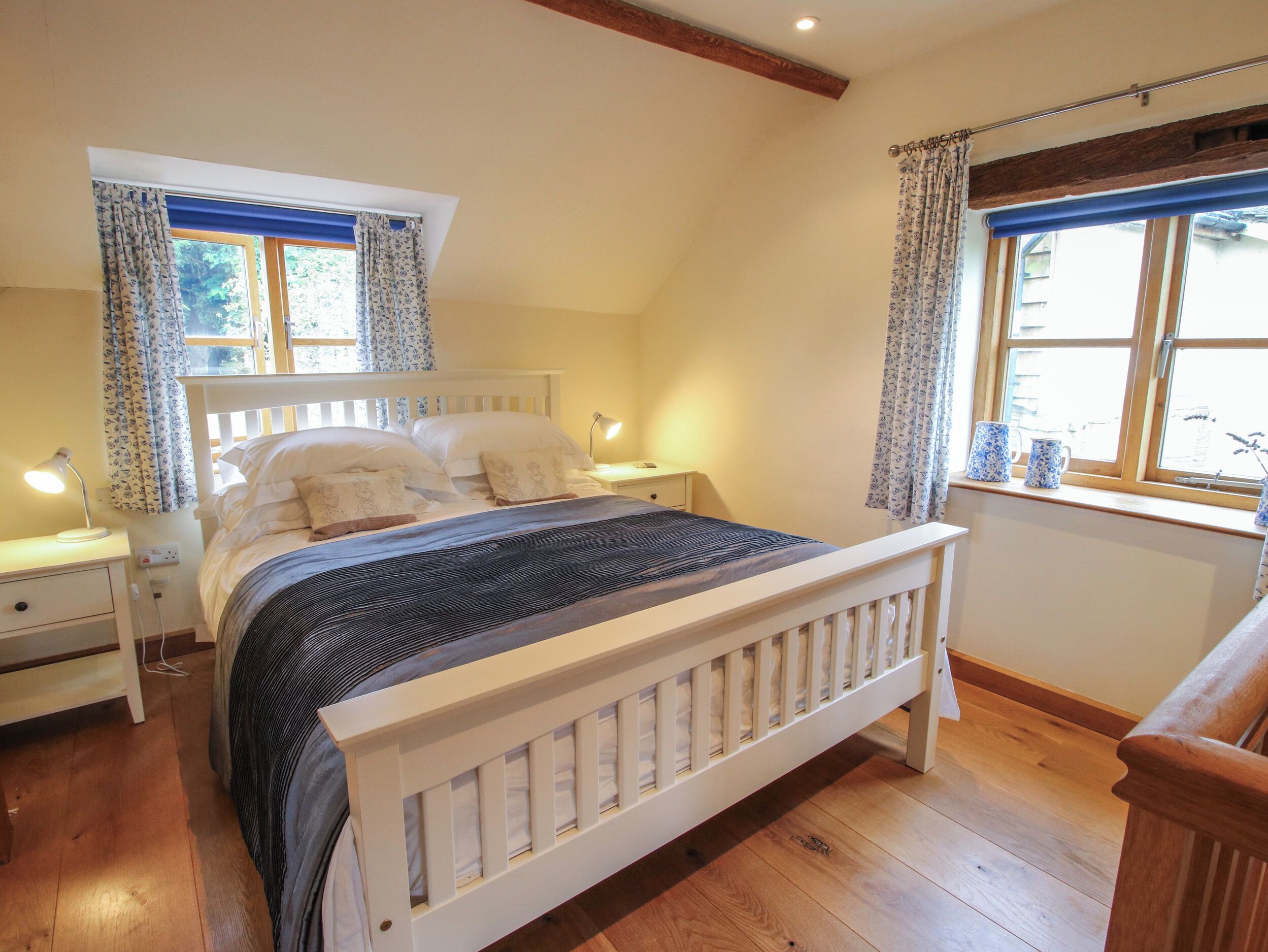 A bedroom with a wooden bed, two bedside tables with lamps, a window with curtains, and a wooden floor.