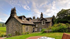 A stone house with a large garden, a wooden deck, and a table with a book. - Riverbank At Stepping Stones (Ambleside)