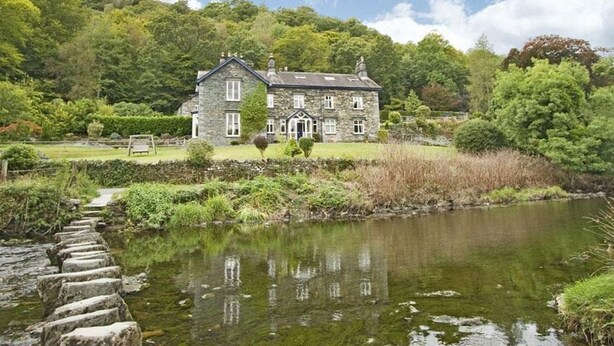 A stone house with a pond in front, surrounded by lush greenery and trees. - Riverbank At Stepping Stones (Ambleside)
