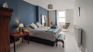 A bedroom with a bed, bedside table, lamp, chair, and a view of the beach through a window. - Tonnau Yr Oleu or Waves of Light (Barmouth)