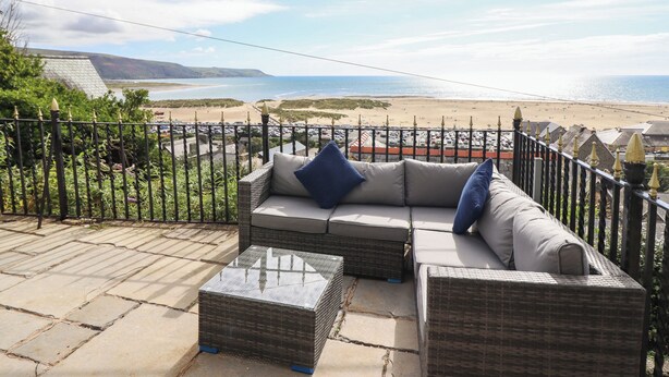 A patio with wicker furniture and a glass table, overlooking a beach with a fence in the foreground. - Tonnau Yr Oleu or Waves of Light (Barmouth)