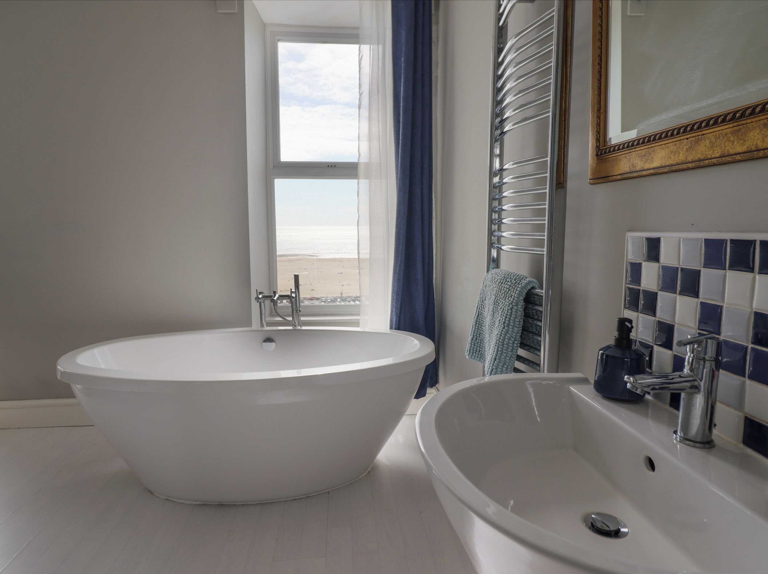 Une salle de bain avec une baignoire sur pieds, un lavabo avec un miroir et une vue sur la plage par une fenêtre.