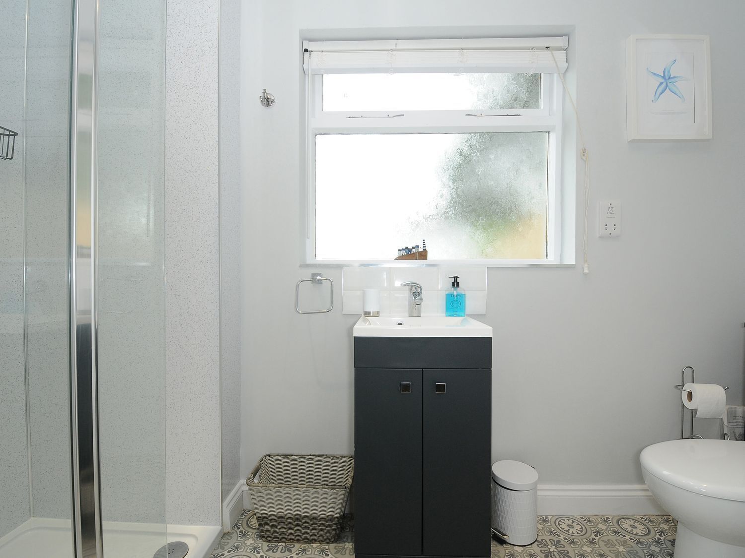 A bathroom with a shower, a sink with a window above, a toilet, and a small trash bin.