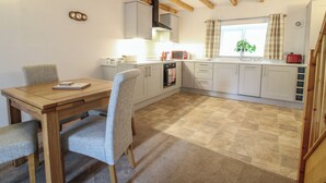 A compact kitchen with white cabinets, a wooden dining table, and a window with curtains. - Ascot (Wrexham)