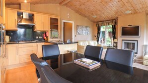 Eine moderne Küche mit Holzschränken und einer Edelstahl-Dunstabzugshaube. Ein Essbereich mit Glastisch und schwarzen Stühlen ist ebenfalls vorhanden. Im Wohnzimmer sind eine Couch und ein Fernseher zu sehen.