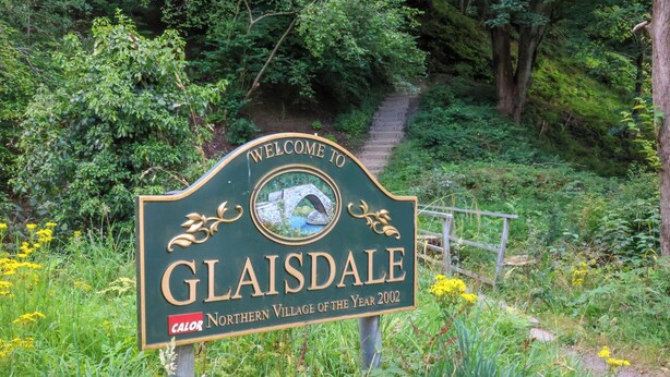 Welkomstbord voor Glaisdale, Noordelijk Dorp van het Jaar 2002, omringd door weelderige begroeiing en een pad.