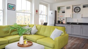 A modern living room with a green sofa, a small table with a plant, and a kitchen area with grey cabinets and a clock. - Harbourside Apartment (Pwllheli)