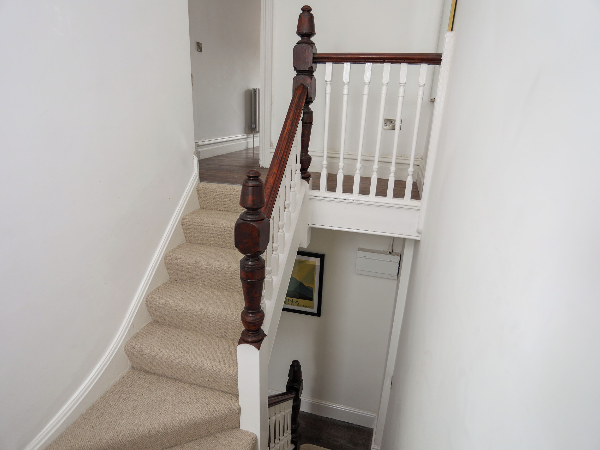Un escalier avec une main courante en bois et des balustres blanches, des marches recouvertes de moquette et un petit tableau encadré accroché au mur.