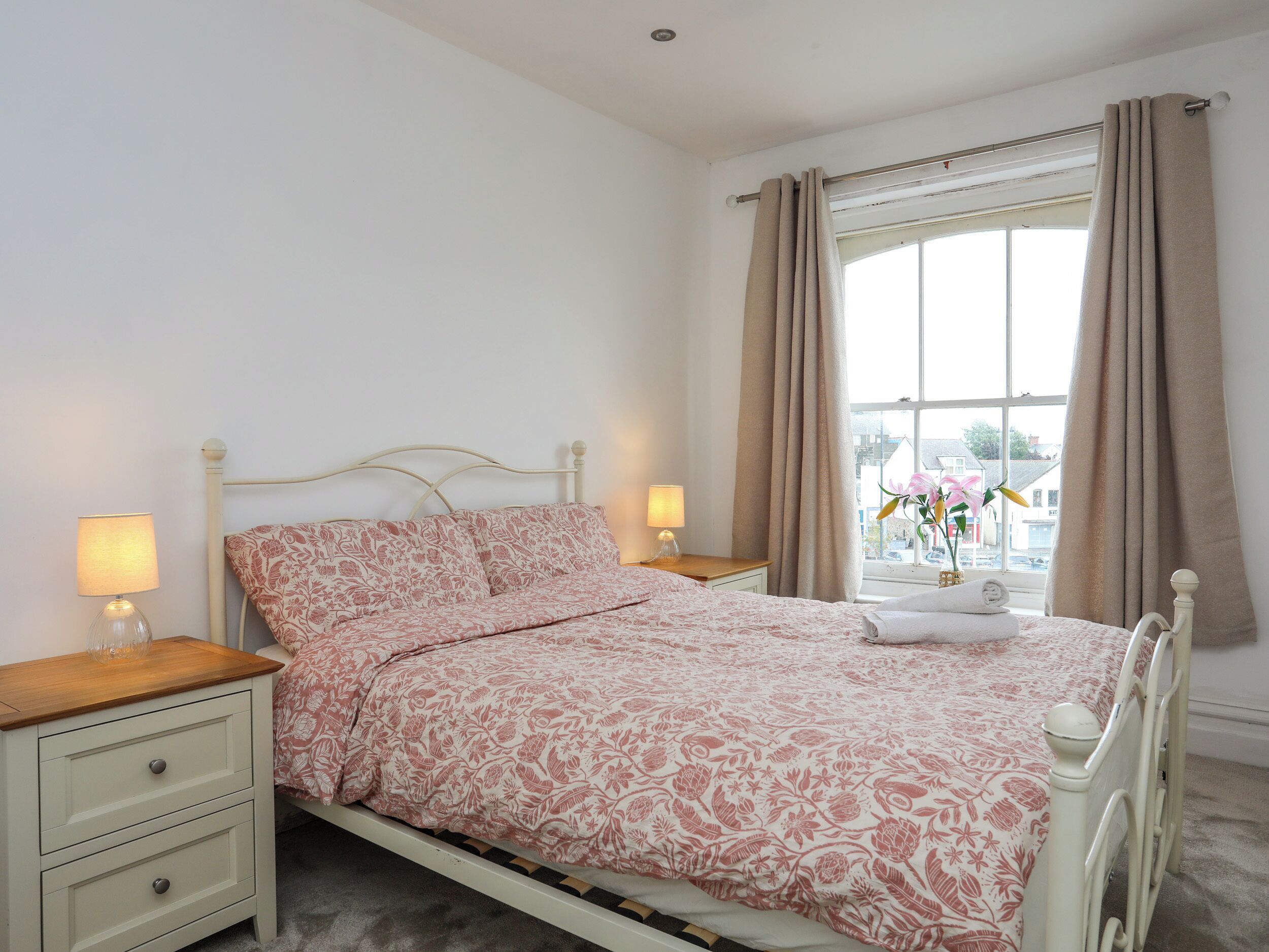 A bedroom with a bed, two bedside tables with lamps, a window with curtains, and a view of the outside.