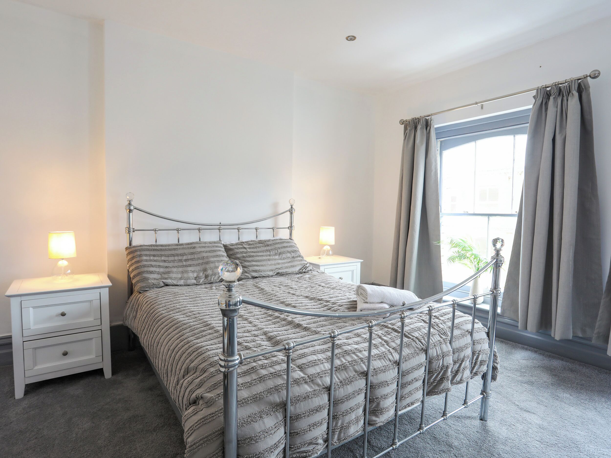 A bedroom with a metal-framed bed, two bedside tables with lamps, a window with curtains, and a view of greenery outside.