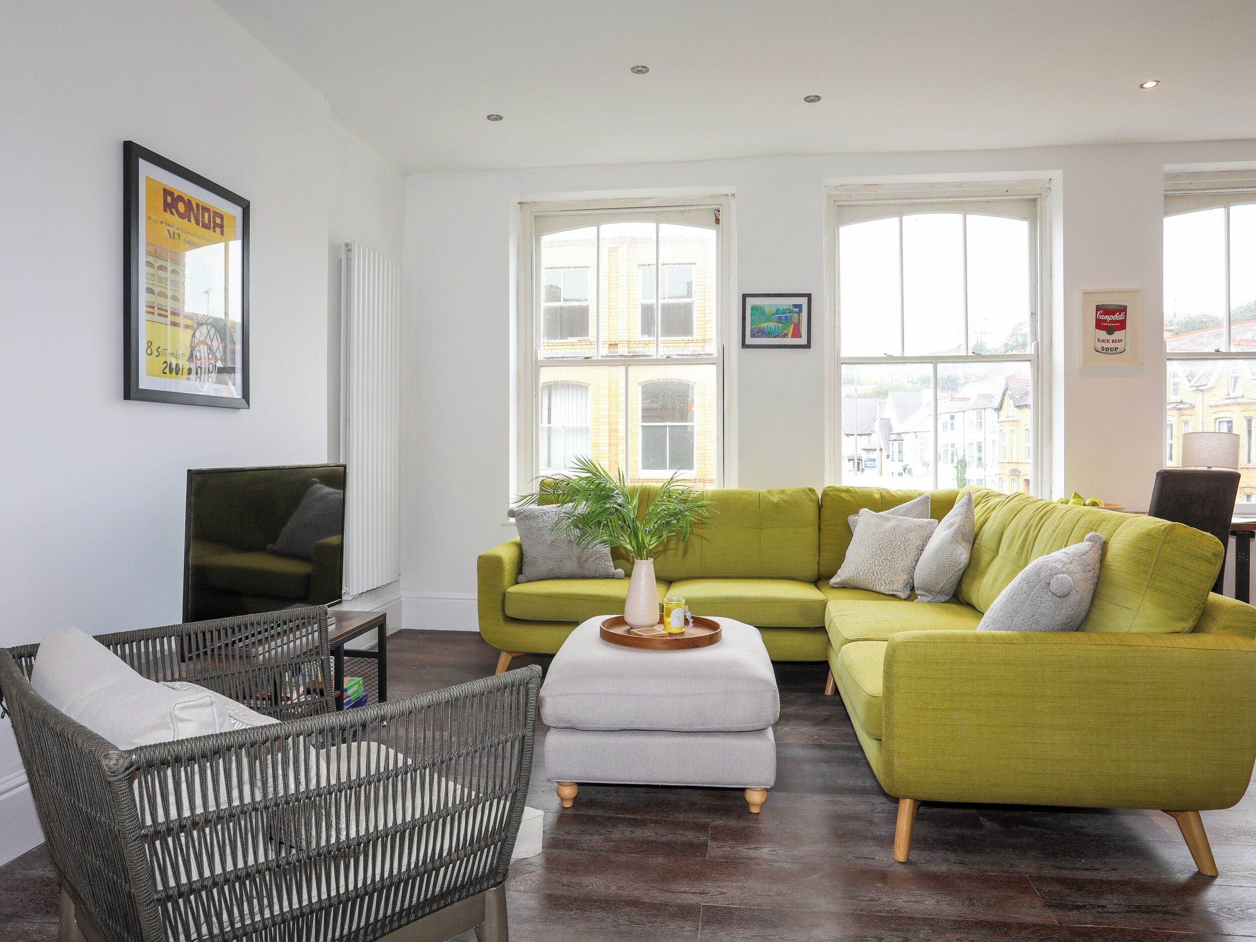 A modern living room with a green sofa, a wicker chair, a white ottoman, and a television.