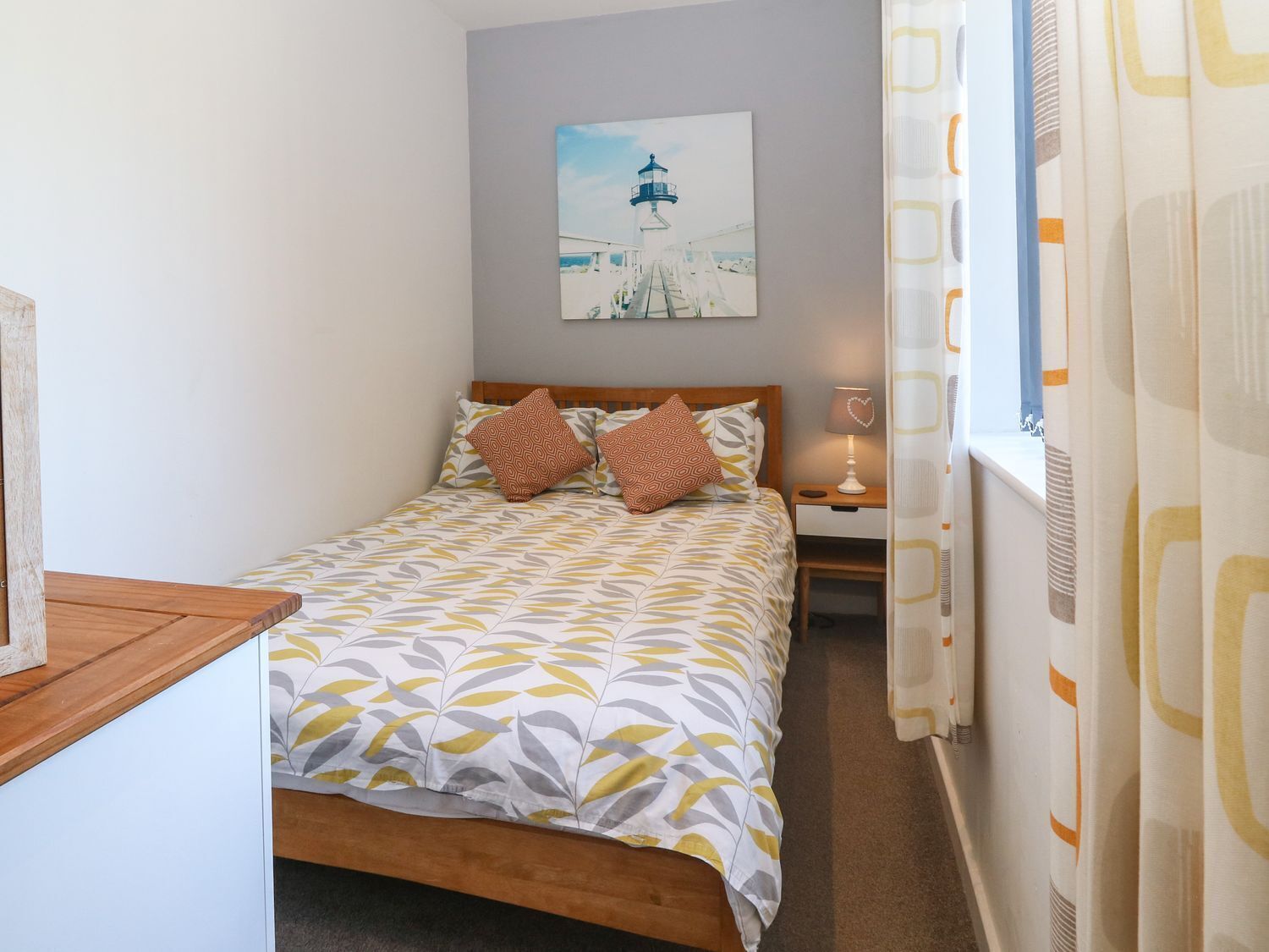 A neatly arranged bedroom with a bed, a bedside table with a lamp, a picture on the wall, and a window with curtains.