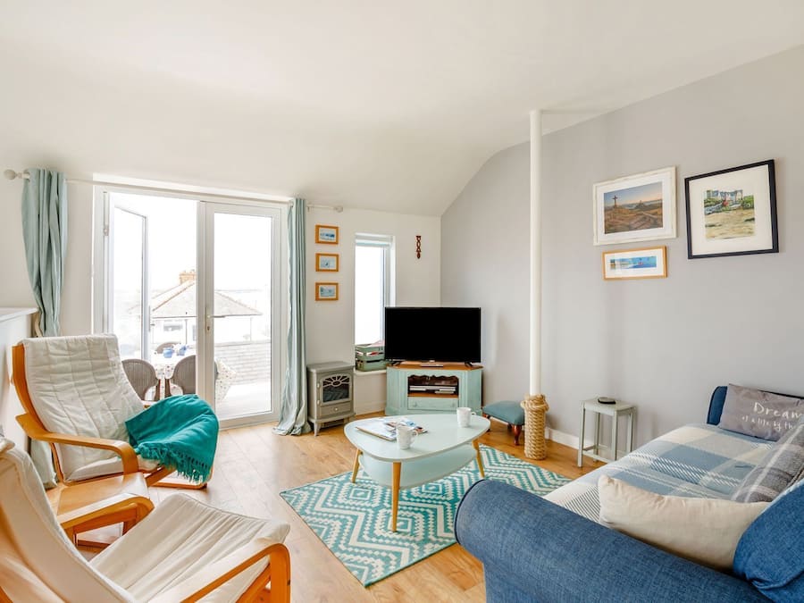 Een gezellige woonkamer met een bank, fauteuil en een televisie. Er is een raam met gordijnen, een klein tafeltje met een kopje, en een vloerkleed met een patroon.