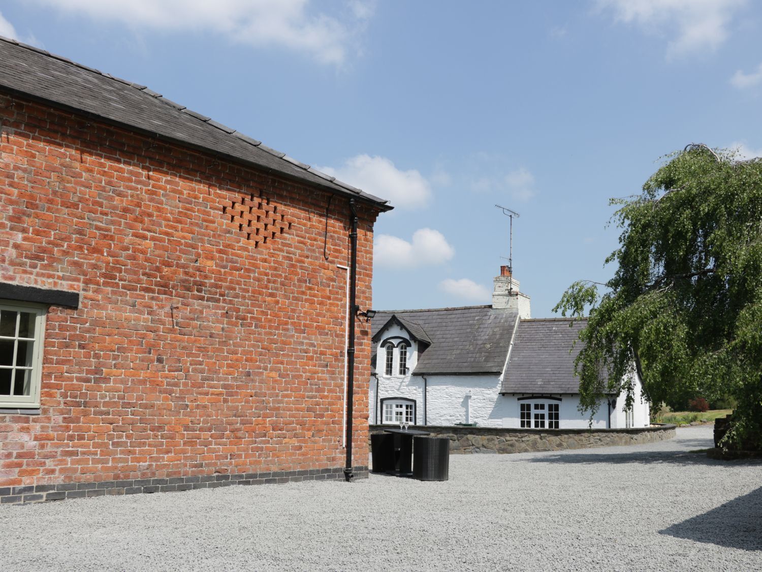 A red brick building with a gravel driveway and a white house with a grey roof in the background.