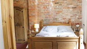 A wooden bed with a headboard, a nightstand with a lamp, a mirror, and a wooden wardrobe. - Woodside Cottage (Newtown)