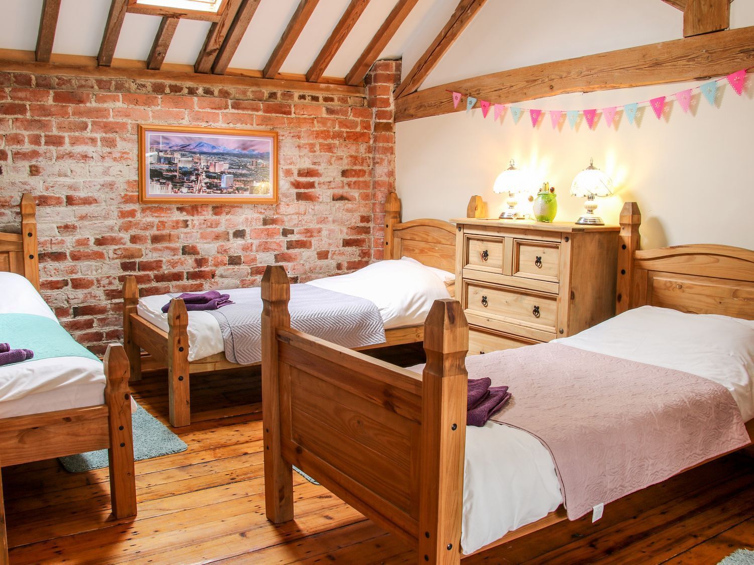 A rustic room with three wooden beds, a brick wall, and a framed cityscape picture.