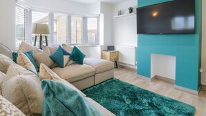 A modern living room with a sofa, a teal accent wall, a flat-screen TV, and a view through large windows. - Jasmine Cottage (Herne Bay)